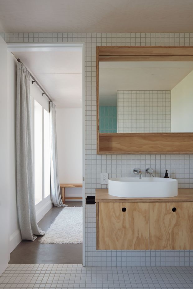 A bright, minimalist bathroom with a white vessel basin set on a natural timber vanity, framed timber mirror, and pale square tiled walls and floor. A doorway opens to an adjacent light-filled space with full-height windows, sheer curtains, and a soft rug. The bathroom features clean lines, restrained detailing, and warm timber accents.
