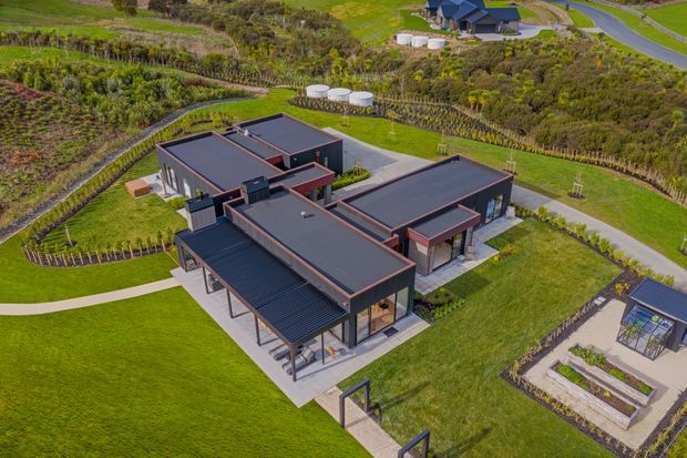 Aerial view of a contemporary single-storey home arranged in several connected wings around landscaped outdoor spaces. The building has dark flat roofs, charcoal cladding, extensive glazing, covered outdoor terraces, and warm timber or reddish-brown trim. It sits within a carefully landscaped rural or semi-rural property with lawns, planted borders, paths, raised garden beds, and nearby outbuildings.