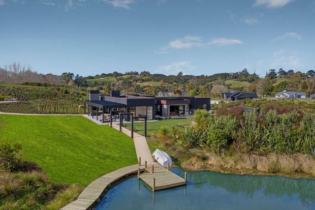 Wide exterior view of a low, modern single-storey residence set within a landscaped site beside a pond. The home has dark horizontal cladding, extensive glazing, a flat roof, covered outdoor areas, and a long walkway leading from the house toward a curved timber deck and small watercraft dock. Rolling green hills and established planting surround the property.