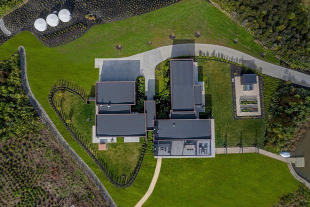 Aerial top-down view of a large modern residential property set within expansive landscaped grounds. Multiple connected flat-roofed building volumes, paved paths, curved driveway, lawns, planted garden areas, and a small detached outbuilding are visible.