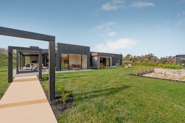 Wide exterior view of a low-rise contemporary compound set within a landscaped grassy site. Dark, rectilinear pavilion-like buildings with large glazed openings are connected by a covered timber or steel-framed walkway. A pale concrete path crosses the lawn, with garden beds and a partially visible outdoor seating area. The image is captured in bright daylight with a clear blue sky.