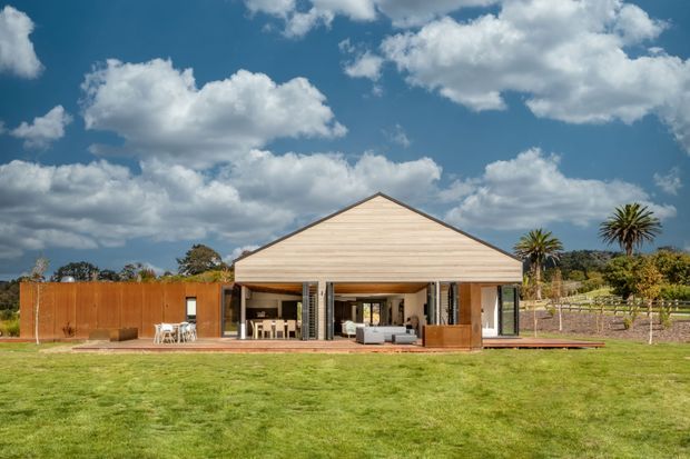 Single-storey contemporary house viewed from the lawn, with a broad gabled timber-clad facade, deep covered outdoor living area, extensive openings, warm timber screening, and a landscaped rural setting with palms and planted rows.
