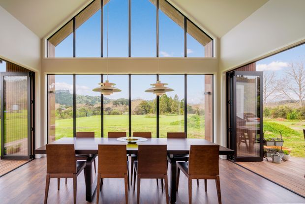 Bright, spacious dining room with a long timber dining table and matching wooden chairs positioned in front of expansive floor-to-ceiling glazing. The room has a dramatic pitched ceiling with clerestory windows, warm timber flooring, slim dark window frames, and pendant lights. Open glazed doors connect the dining space to a landscaped rural garden and lawn.