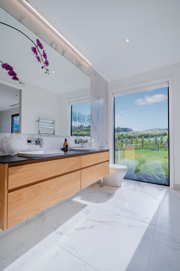 Bright contemporary bathroom with a floating timber vanity, white stone or marble-look benchtop and splashback, integrated basin, wall-mounted tapware, heated towel rail, toilet, and large-format marble-look floor tiles. A full-height glazed door or window provides views to a green rural landscape, while a large mirror and recessed ceiling lighting enhance the airy space.