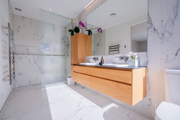 Bright contemporary bathroom with full-height white marble-look wall and floor tiles, a floating timber double vanity with white basins and dark benchtop, large mirrored cabinetry, wall-mounted towel rail, and a toilet. Purple orchids add a small decorative accent.