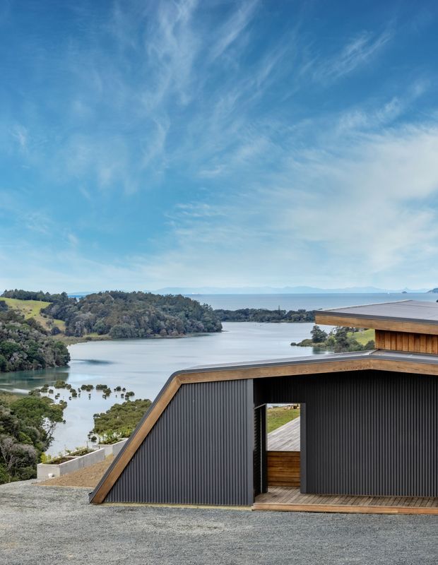 Contemporary dark-clad building with angular sloping roof forms and warm timber trim, positioned above a tranquil waterway with wooded islands and distant hills. The image is an exterior architectural view dominated by the building facade and expansive waterfront setting.