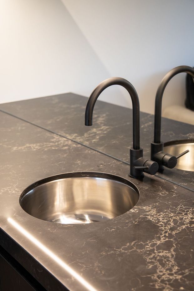 Close-up of a compact kitchen or wet-bar sink set into a dark, veined stone benchtop. The round stainless-steel undermount basin is paired with a matte black gooseneck tap and separate control, with a mirrored reflection visible at the right.