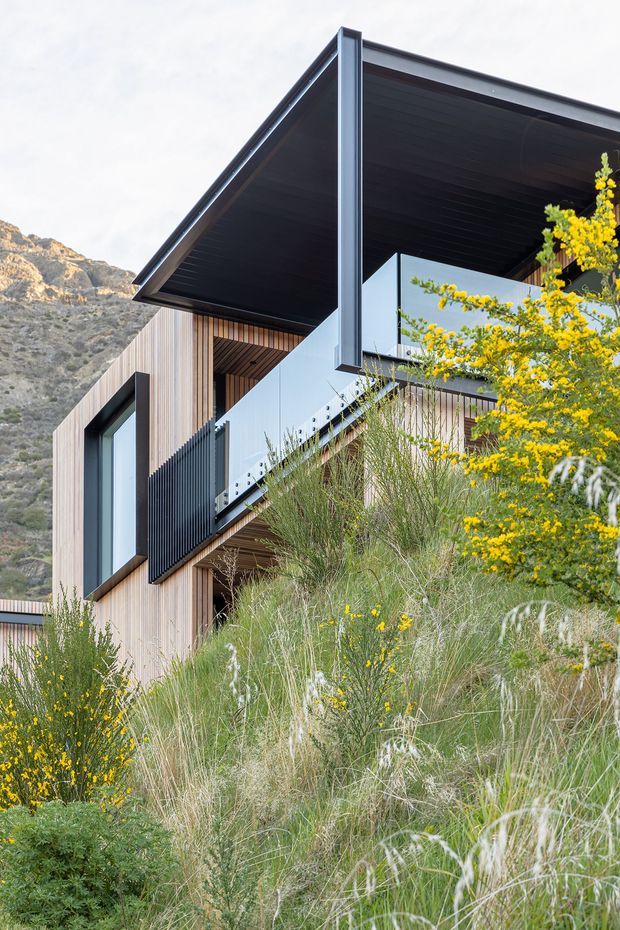 Exterior architectural photograph of a contemporary multi-level house set into a steep, mountainous landscape. The building features vertical timber cladding, dark metal roof planes and soffits, expansive glazing, projecting cantilevered forms, and a dark vertical slatted balcony screen. Native grasses and flowering shrubs occupy the foreground.