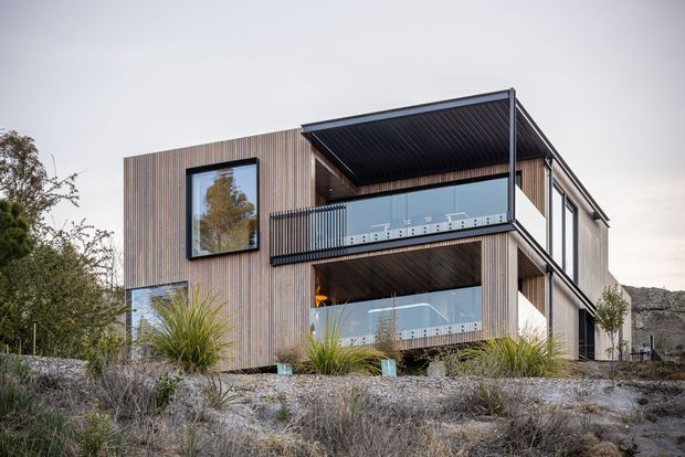 A contemporary multi-level residential house viewed from the exterior, featuring vertically clad timber-look walls, expansive glazing, projecting balconies with transparent balustrades, and a large covered upper terrace. The home sits on a landscaped, rocky site with native-style planting.