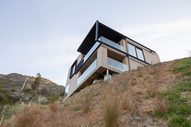 A modern multi-level house perched on a steep, rugged hillside. The building features extensive glazing, black metal framing, projecting balconies with clear glass balustrades, and vertical timber cladding beneath a dramatic angular roof overhang. Native-looking grasses and rocky terrain surround the elevated structure.