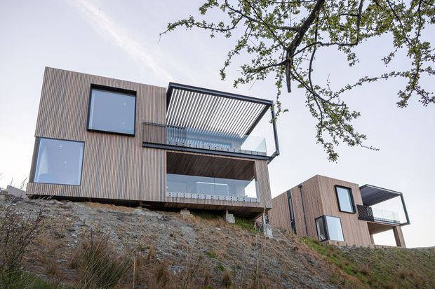 Exterior view of a contemporary multi-level residential building composed of two timber-clad volumes set on a steep rocky hillside. The homes feature large glazed windows, projecting balconies, and prominent slatted roof or privacy screens, with elevated construction above the sloping site.