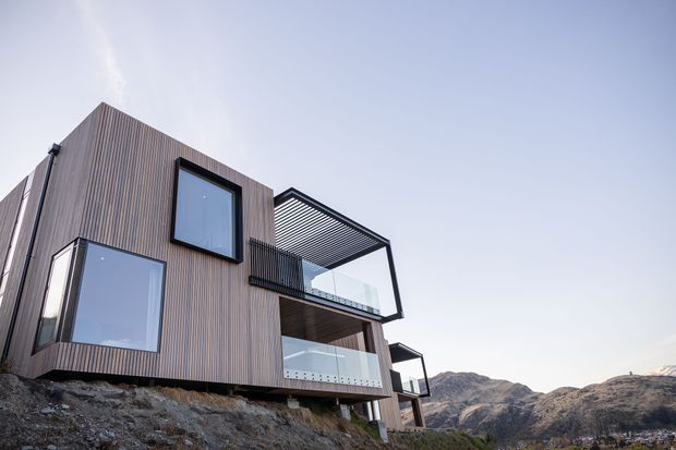 Exterior architectural photograph of a contemporary multi-level house perched on a steep rocky hillside. The building has vertically ribbed timber-toned cladding, large dark-framed windows, projecting glazed balconies, and a striking open pergola-like roof structure. Rugged mountainous terrain is visible behind the dwelling.