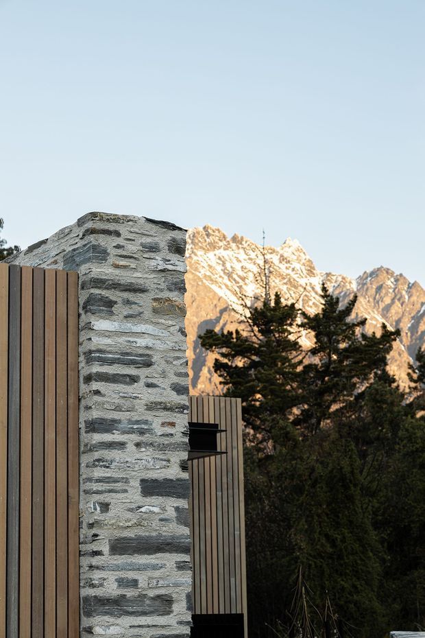 Vertical exterior architectural photograph showing a textured stacked-stone wall framed by vertical dark timber cladding and a partially visible projecting structure. Dense alpine vegetation and dramatic snow-capped mountain peaks form the background in warm low-angle light.