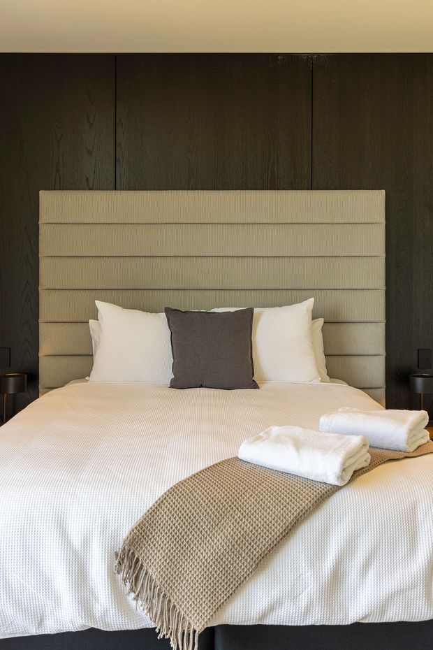 A warmly lit bedroom centered on a neatly made bed with a tall, horizontally channelled upholstered headboard. Dark vertical timber wall panelling forms the backdrop, with small dark bedside tables and lamps on either side. The bed is dressed in light cream-white textured bedding, layered with white pillows, a dark grey accent cushion, folded towels, and a taupe knitted throw.