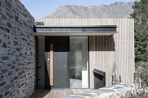 Exterior view of a compact contemporary mountain dwelling, with a dark recessed entry framed by vertical timber cladding, a low flat roof, and a rugged natural-stone wall. A timber deck and stone-paved foreground lead toward the entrance, with dramatic alpine mountains visible behind the building.