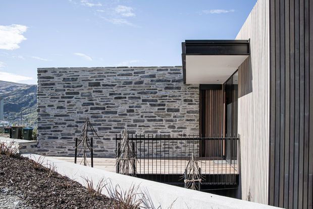A modern residential exterior on a sloped site, combining a long grey stone masonry wall with vertically ribbed dark timber or metal cladding. A recessed entry is sheltered by a projecting flat canopy, with black metal balustrades and a light concrete walkway. Mountainous terrain is visible in the background.