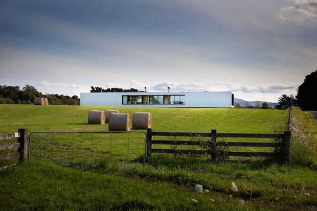 A long, low-slung white contemporary house set in an expansive rural paddock, viewed from behind a timber farm fence. The building has a simple rectangular form, flat roof, crisp white walls, and a broad horizontal glazed opening. Hay bales and open pasture reinforce the rural setting, with distant wooded hills under an overcast sky.