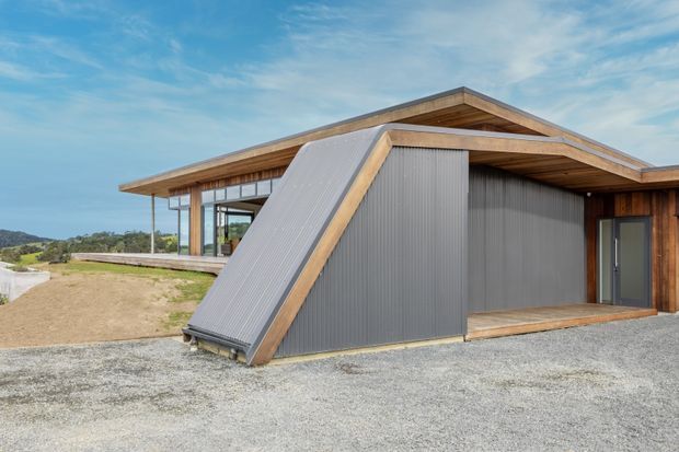 Exterior view of a contemporary single-storey building with a prominent asymmetrical gable roof and a dramatic sloping corrugated-metal-clad wall. The structure combines warm timber cladding and exposed timber roof framing with extensive floor-to-ceiling glazing, set in an open gravelled landscape.