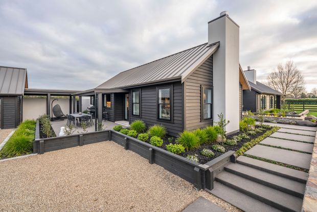 Exterior view of a contemporary single-storey home with dark horizontal cladding, a pitched metal roof, prominent white chimney volumes, and extensive landscaped garden beds. The property includes a gravel forecourt, stepped paved walkway, raised dark-edged planters, outdoor dining area, and covered outdoor connection between building volumes.