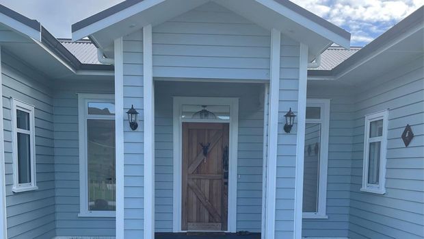 Front exterior entry of a single-storey home with pale blue horizontal weatherboard cladding, white trim, a prominent gabled porch canopy supported by square posts, and a rustic timber plank front door. Symmetrical wall lanterns and white-framed windows flank the entrance.