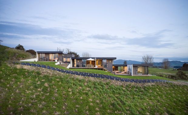 Wide exterior view of a low-profile contemporary residence arranged across a gently sloping grassy site. The house comprises several connected pavilion-like volumes with flat roofs, extensive glazing, dark cladding and warm timber accents. A paved driveway runs across the foreground, with open rural or semi-rural landscape and distant hills beyond.