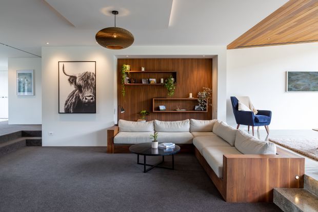 A refined contemporary living room with a large cream sectional sofa, dark round coffee table, and built-in walnut-toned shelving and cabinetry. The room features dark carpet, white walls, a sculptural pendant light, recessed linear lighting, timber ceiling detailing, artwork, plants, and a blue lounge chair. Steps lead toward an adjacent area at the left, while a raised seating nook with timber ceiling lining is visible on the right.