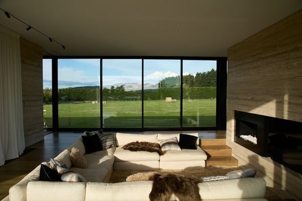 A spacious contemporary living room with a large cream sectional sofa arranged around a broad built-in fireplace. Full-height black-framed glazing opens views to a landscaped lawn and distant hills, while warm horizontal timber wall cladding and dark timber flooring create a natural, refined interior. Track lighting and soft neutral furnishings complete the calm, luxurious setting.