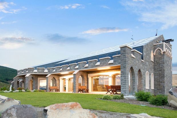 Exterior view of a substantial stone-clad building with a long, low wing and crenellated parapet, connected to a taller Gothic-inspired gabled volume. Large glazed openings and covered outdoor seating face a manicured lawn, with rocky terrain and wooded hills in the background. The building has a distinctive castellated, masonry-heavy character and a bright metal pitched roof.