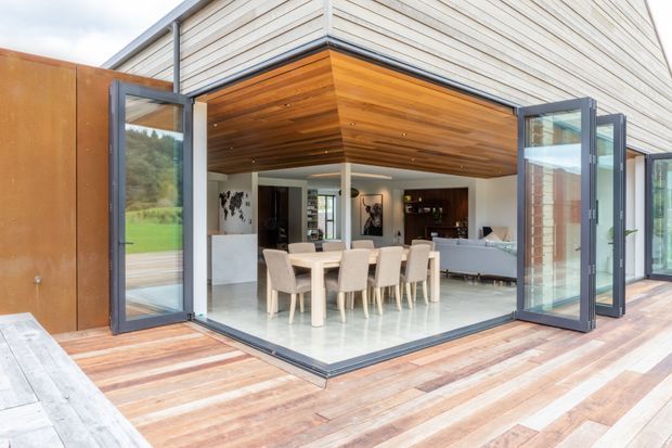 A contemporary open-plan dining and living area opening onto a timber deck through fully glazed, folding corner doors. The space features a large dining table, pale upholstered chairs, white walls and flooring, a dark kitchen or bar joinery wall, and a warm timber-lined recessed ceiling beneath a light horizontal-clad exterior volume.