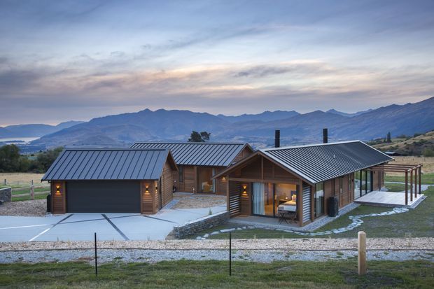 A single-storey rural mountain dwelling composed of several connected timber-clad pavilions with dark standing-seam metal gable roofs. The home sits on a broad gravelled and paved site, with large glazed openings and a covered outdoor terrace, against a dramatic mountain backdrop at dusk.