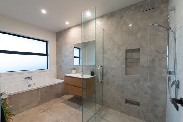 Modern bathroom featuring a freestanding bathtub beneath a large black-framed window, a timber-vanity with basin and mirror, and a spacious frameless glass shower enclosure. The shower and surrounding walls are clad in textured grey stone-look tile, with warm neutral floor tiles and recessed lighting.