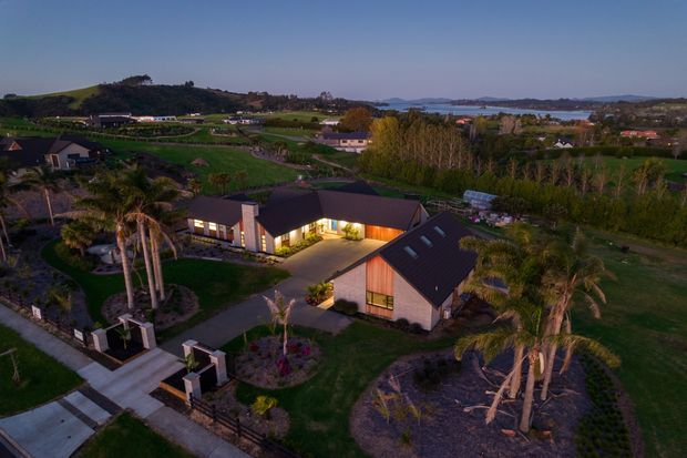 Aerial dusk view of a modern rural residential property with several dark pitched-roof buildings, illuminated interiors, landscaped gardens, palm trees, and a paved driveway. Rolling green farmland and a distant coastal or waterside landscape are visible beyond the property.