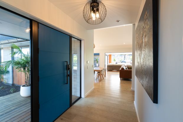 A bright contemporary residential entry hall with a large deep-blue pivot front door, full-height black-framed glazing, warm timber flooring, white walls, and a black wire pendant light. The hall opens toward a dining and living area, with a planted courtyard visible through the glazing and a large textured wall artwork along the right side.