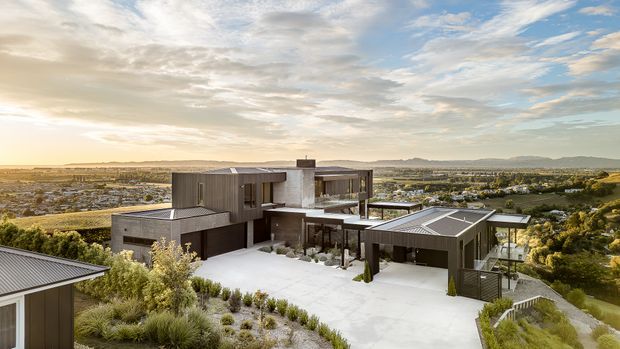 A large contemporary multi-level residence viewed from above, set on an elevated landscaped site overlooking a suburban valley and distant hills. The home features strong horizontal and rectilinear forms, dark vertical cladding, extensive glazing, broad flat roof planes, rooftop solar panels, and a spacious pale paved forecourt. Native-style planting surrounds the building, with a separate garage or outbuilding connected by the driveway.