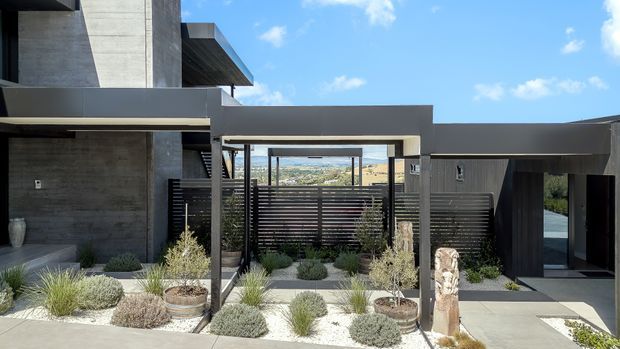 Modern multi-level residential exterior with dark charcoal and exposed concrete volumes, flat rooflines, deep overhangs, and a covered entry courtyard. Horizontal slatted screening, drought-tolerant ornamental planting, pale gravel, and concrete paving define the landscaped forecourt. The open central view reveals a distant hilly landscape.