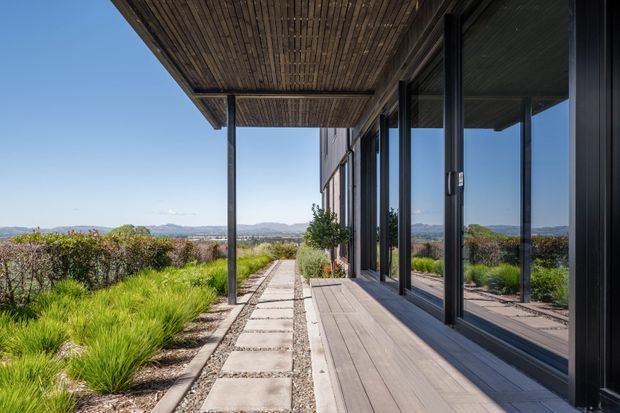 A contemporary glazed building edge with a long covered outdoor terrace or verandah. Full-height dark-framed glass doors reflect the terrace, while a timber-lined soffit and timber decking create a sheltered transition to a concrete-paver walkway. Native-style landscaping and distant hills are visible beyond.