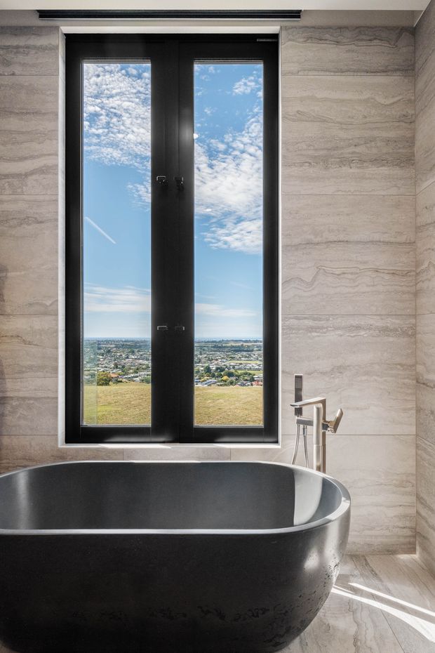 A refined bathroom centered on a freestanding matte-black bathtub positioned in front of tall black-framed windows. The walls and floor are clad in large-format pale beige-grey veined stone, with a minimalist chrome floor-mounted bath tap and hand shower. Bright daylight and a broad elevated landscape view create a dramatic, luxurious atmosphere.