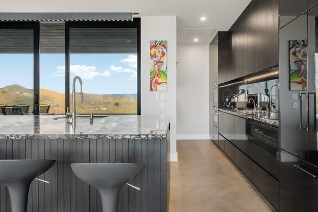 A contemporary galley kitchen with a dark timber-toned wall of integrated cabinetry on the right and a marble-topped island in the foreground. The island features a vertically fluted dark facade and two sculptural grey bar stools. Large black-framed windows provide expansive views of an open hilly landscape, while mirrored cabinetry reflects the sink and kitchen workspace. Colourful artwork adds a small accent beside the windows.