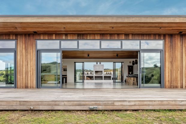 Wide exterior view of a contemporary timber-clad pavilion or house opening onto a broad timber deck. Large sliding glass doors and clerestory glazing reveal a bright open-plan interior with a central fireplace and kitchen or dining area. Rolling green countryside is visible beyond the glazing, while the building features strong horizontal roof eaves and warm vertical timber cladding.