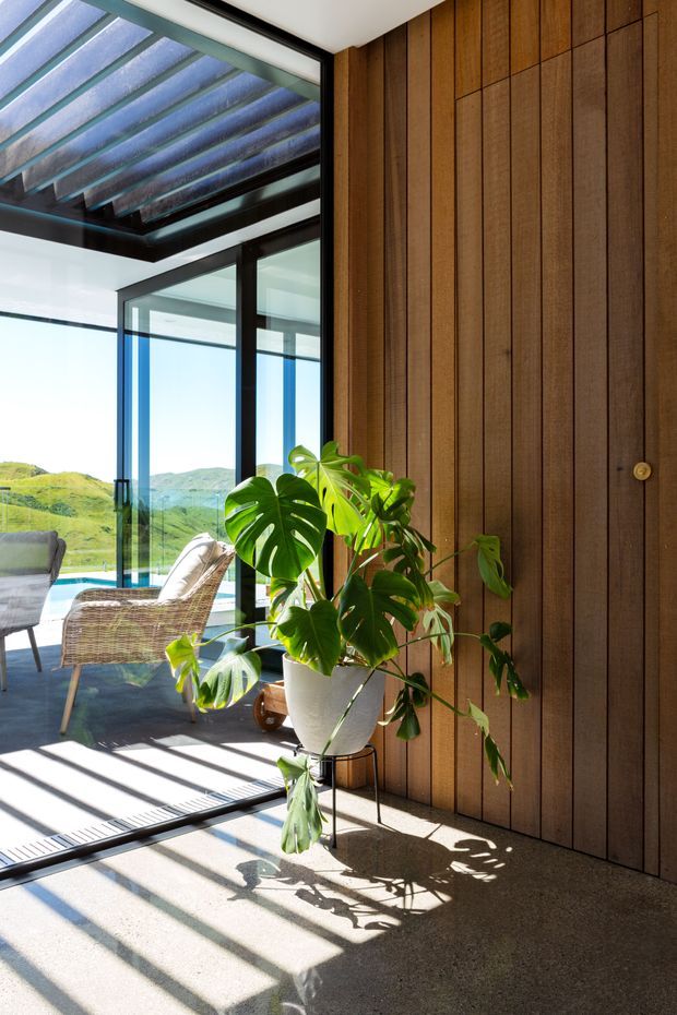 A bright contemporary interior corner with full-height black-framed glazing opening onto a scenic green landscape and pool. The space features a vertical dark timber-panelled wall with concealed joinery, polished concrete flooring, and a large potted monstera plant. Strong sunlight casts linear shadows across the floor, while an adjacent glazed outdoor or living area contains wicker seating and a dining table. A glazed, slatted roof structure is visible overhead.
