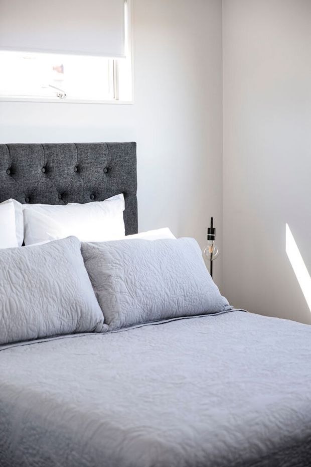 A bright, close-up view of a neatly made bedroom. The bed has a dark grey upholstered, button-tufted headboard, white pillows, and pale grey quilted bedding. White walls and a partially covered window create a clean, minimal setting, with a slim black bedside lamp and strong sunlight casting a geometric patch of light on the wall.