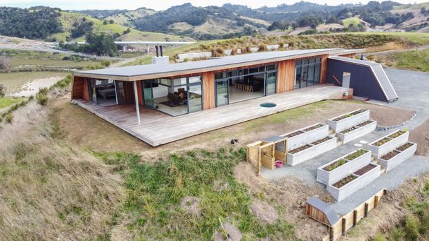 Aerial view of a contemporary single-storey home set on a sloped rural landscape. The long, low-profile dwelling has a flat roof, extensive glazing, timber cladding and a broad timber deck. Raised garden beds and landscaped gravel areas sit in front of the house, with rolling hills and a roadway visible beyond.