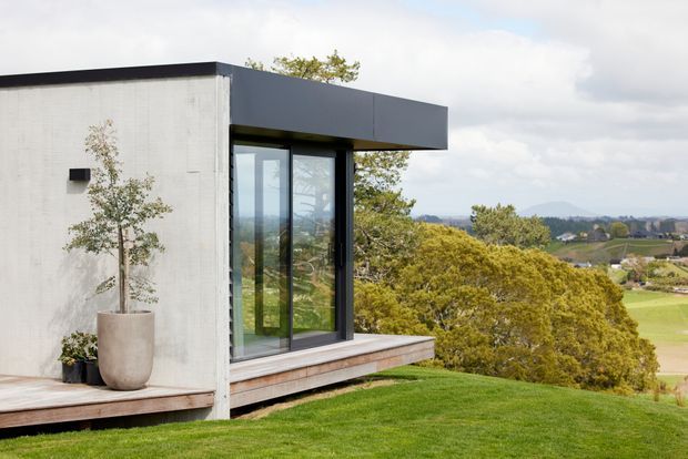 Exterior view of a contemporary flat-roof building with a minimalist concrete or rendered facade, dark projecting roof fascia, and large floor-to-ceiling glazed opening in a dark frame. A timber-clad deck projects from the building, overlooking a landscaped lawn and open rural landscape.