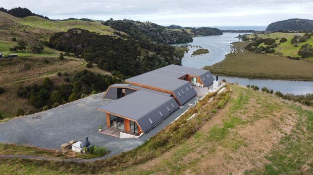 Aerial view of a contemporary multi-wing rural building with dark pitched metal roofs, timber-framed glazed openings, and grey exterior walls, set on a sloping landscaped site beside a winding inlet or estuary. Rolling green hills and water dominate the surrounding landscape.