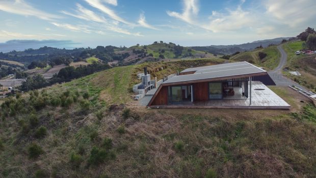 A low-profile modern house set into a gently sloping rural landscape, viewed from above. The home has a broad shallow roof, dark timber-clad walls, extensive glazing, and a large timber deck projecting across the front. Rolling green hills and a winding access road surround the dwelling.