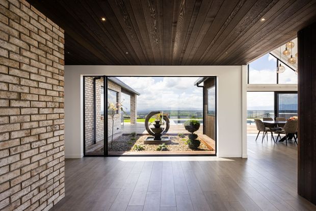A refined contemporary interior opening onto a landscaped courtyard and expansive distant views. The room features a dark timber-lined ceiling with recessed spotlights, pale painted walls, wide timber flooring, an exposed light brick feature wall, and large black-framed glazed doors. A dining area is visible to the right, while the courtyard includes sculptural planting and a water feature or basin.
