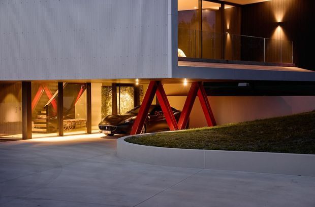 Twilight exterior view of a contemporary multi-level residence with a dramatic cantilevered upper volume, dark horizontal soffits, and a pale vertically ribbed facade. Warm interior lighting reveals a glazed entry and staircase, while bold red angular structural supports frame a parked car beneath the upper level. A curved raised lawn bed and broad concrete driveway occupy the foreground.
