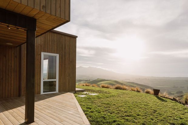 A modern timber-clad building viewed from a covered outdoor deck, with strong vertical wood cladding, exposed timber posts, a timber-lined soffit, and a glazed door. The deck opens onto a grassy elevated landscape with rolling hills and a misty mountain backdrop under a cloudy sky.