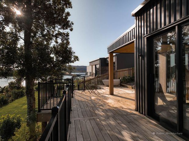 Exterior view of a contemporary multi-level home overlooking a body of water and distant hills. The scene focuses on a broad timber deck with black metal balustrades, dark vertically ribbed cladding, large glazed doors, and a covered outdoor sitting area supported by timber posts. Mature trees and landscaped grounds frame the view.