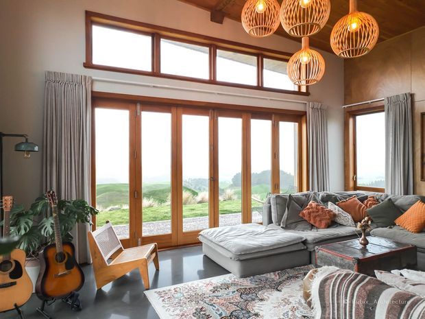 A warm, spacious living room with floor-to-ceiling timber-framed glazing and clerestory windows overlooking a green landscape. The room features a large grey sectional sofa, patterned rug, timber ceiling beams, guitar display, and several woven pendant lights. Draped windows and layered cushions create a relaxed, inviting atmosphere.